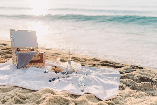 A blanket on the beach with a basket, food, and glasses on it. 