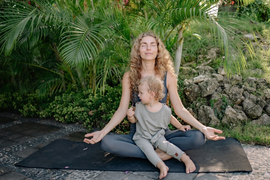 A woman sitting cross-legged on a yoga mat with her toddler in her lap while meditating. 