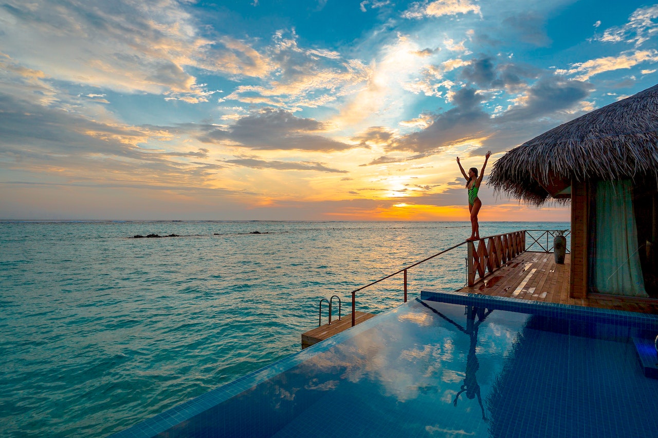 A woman getting ready to dive into the vast blue sea during a sunset with a wooden cottage and infinity pool beside her.
