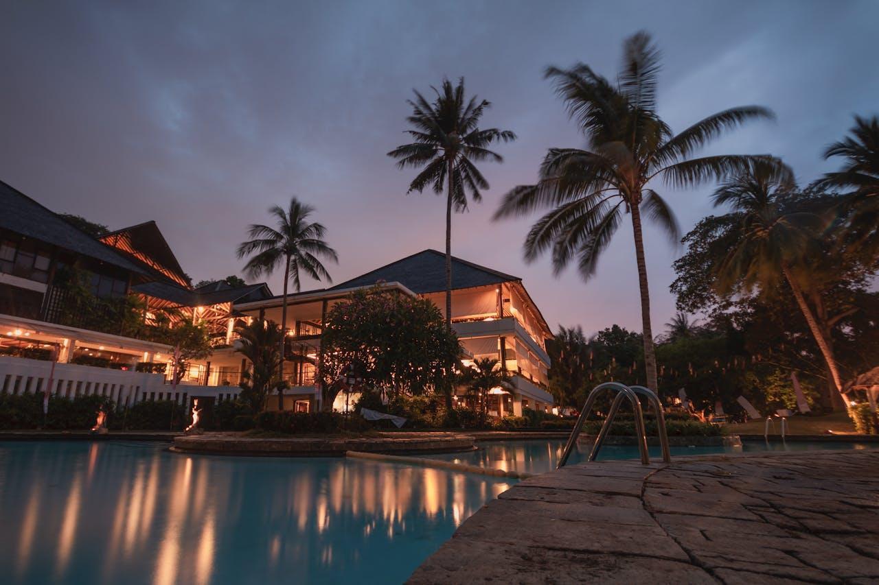 A photo of a swimming pool and a villa in the distance surrounded by palm trees. 