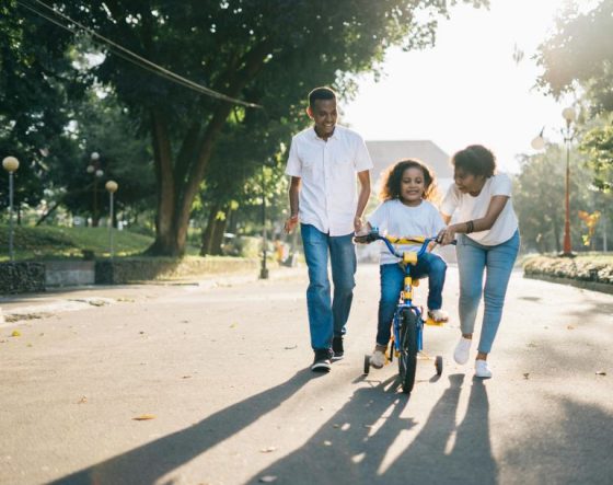 A family helping their child learn to ride a bike in a sunny park.